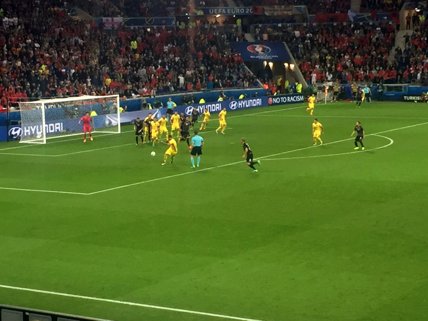 Eine Gruppe von Menschen, die auf einem Feld in einem Stadion Fussball spielen, mit einem Netz auf der linken Seite, beleuchtet von Scheinwerfen von oben, und Zuschauern in den Tribünen hinter Tafeln mit Text.