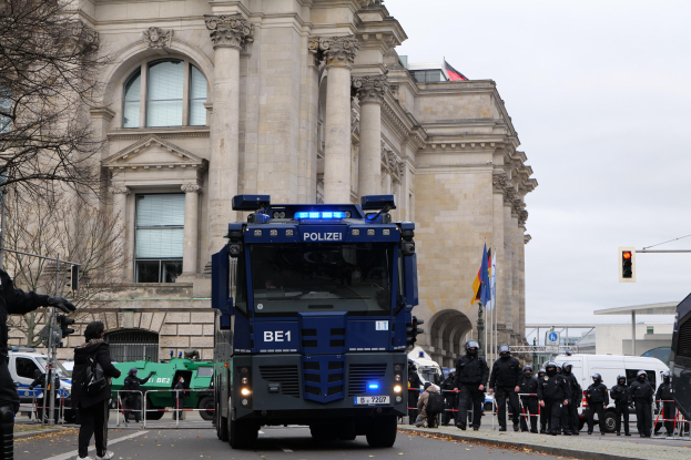 Eine Gruppe von Polizisten steht vor einem großen Gebäude mit Fenstern, Säulen und Bögen, während Fahrzeuge auf der Straße vorbeifahren und eine Person mit einer Kamera auf der linken Seite zu sehen ist. Im Hintergrund sind Bäume, Verkehrsampeln, Fahnenmasten und ein klarer blauer Himmel zu sehen.