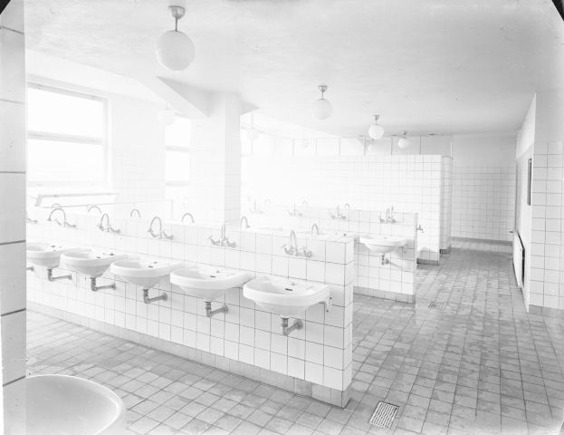 Schwarzes und weißes Foto einer öffentlichen Toilette mit Waschbecken, Wasserhähnen, Wänden, Lampen und Fenstern.