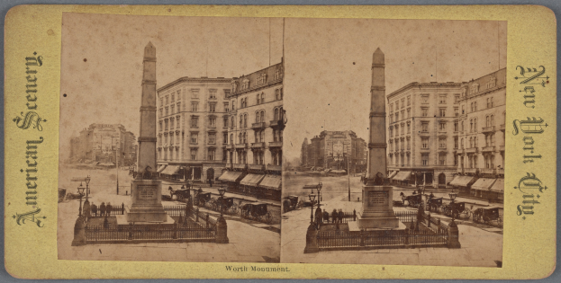 Altes Schwarz-Weiß-Foto einer Stadtstraße mit einem zentralen Denkmal, flankiert von Gebäuden, Straßeneinrichtungsgegenständen, parkenden Autos und Fußgängern auf dem Gehweg unter einem sichtbaren Himmel.