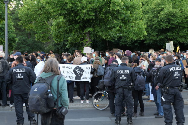 Eine große Gruppe von Menschen steht auf der Straße und hält Schilder und trägt Mützen und Taschen während einer Black Lives Matter Demonstration in Berlin, mit einem Fahrrad im Vordergrund und Bäumen und einem Pfahl im Hintergrund.