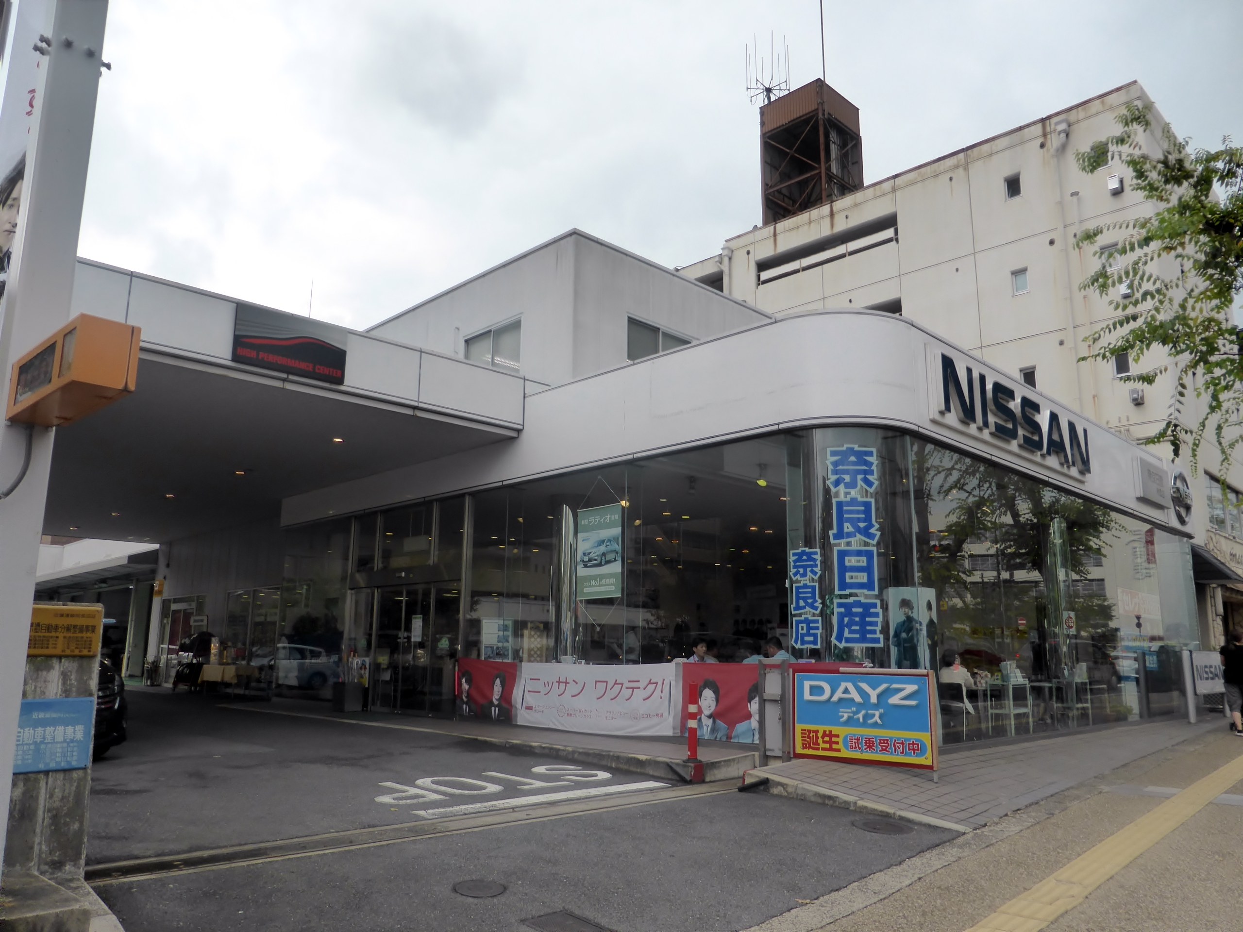 Nissan-Autohaus mit Glaswänden und -fenstern, einem parkenden Auto davor, Schildern mit Text, einem Baum rechts, einer Straße unten und Himmel im Hintergrund.