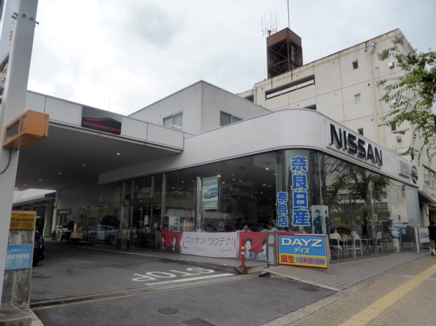 Nissan-Autohaus mit Glaswänden und -fenstern, einem parkenden Auto davor, Schildern mit Text, einem Baum rechts, einer Straße unten und Himmel im Hintergrund.