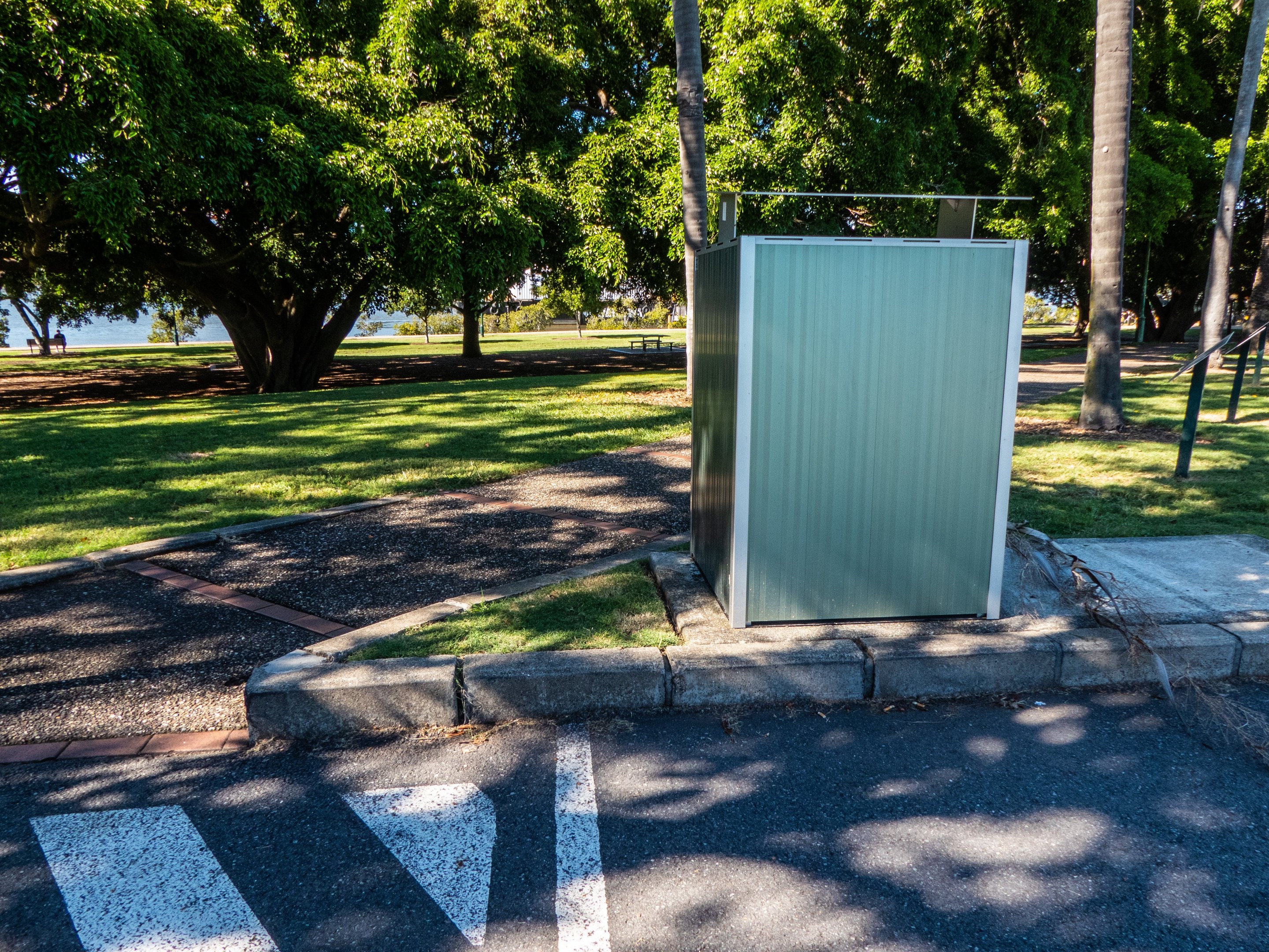 Mobile Toilette am Rande einer Parkstraße in einem Park, umgeben von Bäumen und Gras, mit einem Gewässer und einem klaren blauen Himmel im Hintergrund.
