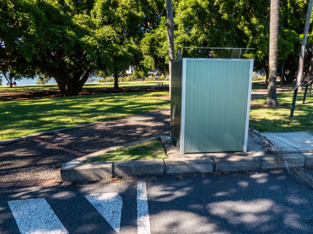 Mobile Toilette am Rande einer Parkstraße in einem Park, umgeben von Bäumen und Gras, mit einem Gewässer und einem klaren blauen Himmel im Hintergrund.