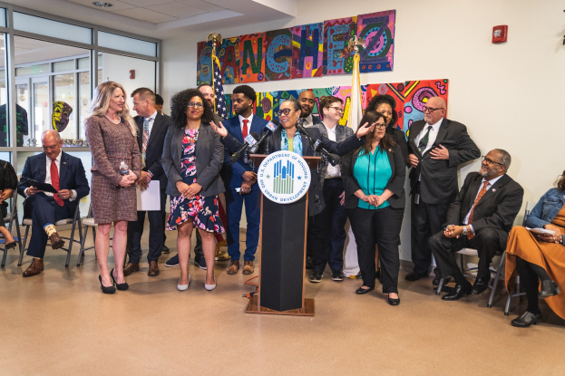 Gruppe von Menschen vor einem Rednerpult mit Mikrofon und einer Tafel mit der Aufschrift "Sancho-Stadtrat tritt dem Stadtrat bei", mit sitzenden und stehenden Teilnehmern, Fahnen, einem Feuerlöscher und Deckenleuchten im Hintergrund.