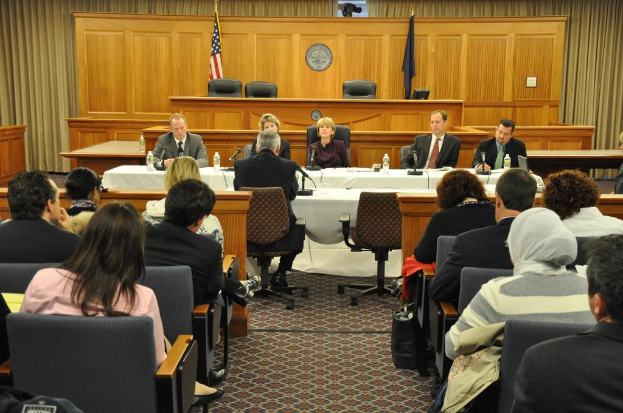 Ein Konferenzraum mit einer Gruppe von Menschen, die sitzen, zwei ausländische Flaggen, ein oranger Schreibtisch mit Wasserflaschen und ein Mikrofon.