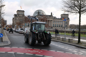 Traktor fährt auf einer von Bäumen und Laternen gesäumten Straße vor dem Reichstaggebäude in Berlin, Deutschland, mit Passanten und Fahrradfahrern auf dem Gehweg, unter einem bewölkten Himmel und dem Gebäude mit Flaggen geschmückt.