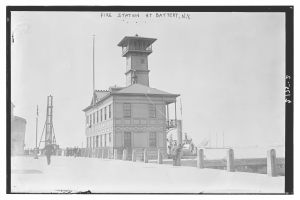 Schwarzes und weißes Foto einer Feuerwache in Battery, New York, mit einem Gebäude, Menschen, Pfählen, einem Zaun, Booten auf dem Wasser und einem bewölkten Himmel.