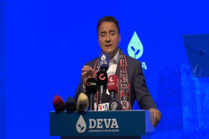 Ein Mann steht an einem Podium mit Mikrofonen, trägt einen Mantel und eine Schärpe und hält ein Schild mit der Aufschrift "deva-Geschäftsführer sagt, es sei keine gute Idee für die Zukunft".