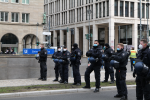 Polizeibeamte in schwarzen Uniformen und Masken vor einem Berliner Gebäude mit Glasfenstern und Säulen, einige halten Helme, mit Laternen, Ampeln, Schildern, einer Statue und anderen Menschen im Hintergrund.