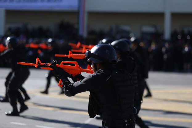 Polizisten in Einsatzausrüstung marschieren mit Waffen die Straße entlang, mit einem Gebäude und einer Fahne im Hintergrund.