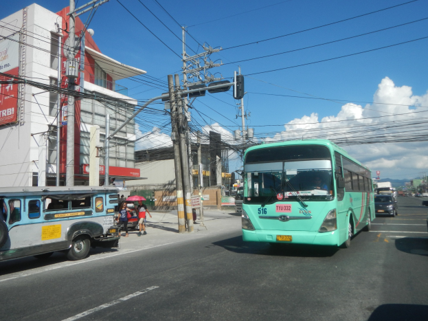 Ein grüner Bus fährt auf einer Straße mit hohen Gebäuden entlang, mit Fußgängern mit Regenschirmen auf dem Gehweg und Strommasten mit Drähten entlang der Straße unter einem bewölkten Himmel.