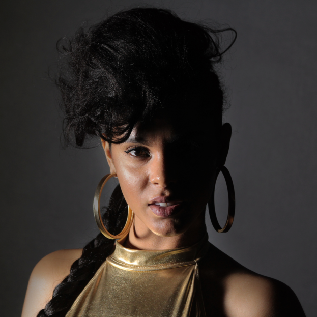 Eine Frau in einem goldenen Kleid und großen Ohrringen steht selbstbewusst vor einer Wand, ihr glatter Bob und natürliches Make-up unterstreichen ihren entschlossenen Gesichtsausdruck, während sie direkt in die Kamera schaut.