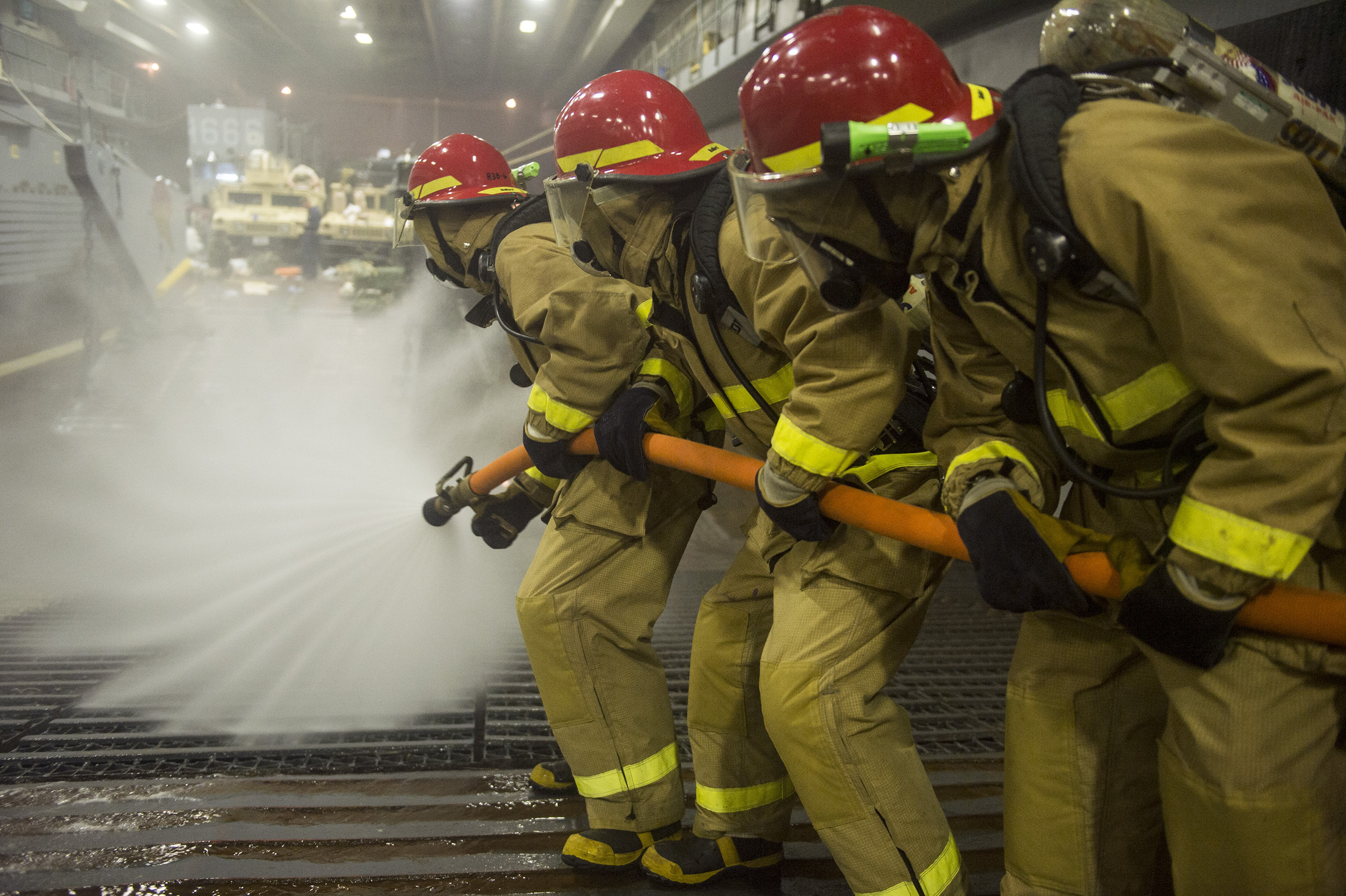 Eine Gruppe von Feuerwehrleuten in Helmen und Handschuhen hält Schläuche und spritzt Wasser auf ein Feuerwehrauto, mit verschiedenen Gegenständen und einem Boden im Hintergrund.