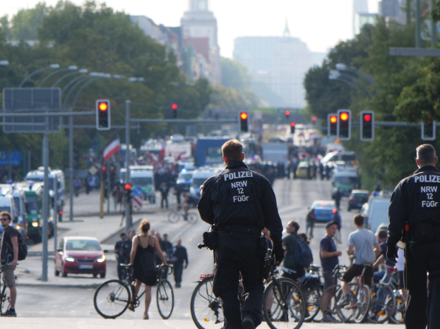 Zwei Polizeibeamte neben einer Gruppe von Radfahrern auf einer Straße mit Fahrzeugen, Verkehrsampeln, Bäumen, Gebäuden und einem klaren Himmel im Hintergrund.
