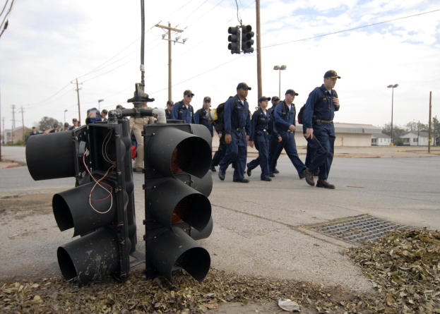Eine Gruppe von Polizisten in Uniform und Mützen, die eine Straße mit Strommasten und Drähten entlanggehen, mit Gebäuden, Bäumen und einem klaren blauen Himmel im Hintergrund.