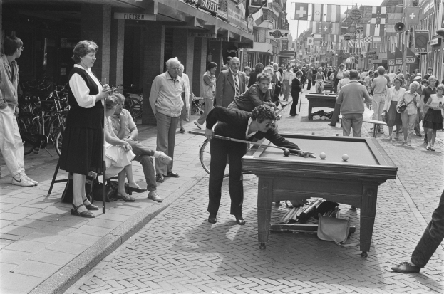 Gruppe von Menschen, die Pool auf einer Stra├če mit Fahrr├Ądern, Geb├Ąuden, Schildern, Fahnen und Masten im Hintergrund spielen.