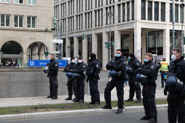 Eine Gruppe von Polizisten in schwarzen Uniformen und Masken steht vor einem Glasfenstergebäude mit Säulen in Berlin, Deutschland, einige halten Helme, mit Laternen, Verkehrssignalen, Texttafeln, einer Statue, anderen Menschen und grasbewachsenem Boden im Hintergrund.