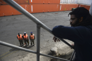 Ein Mann in legerer Kleidung steht auf einem Metallgeländer neben einer Gruppe von Menschen mit orangen Jacken und Kappen, mit Containern und einer Wand im Hintergrund am Hafen von Los Angeles.