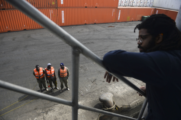 Ein Mann in legerer Kleidung steht auf einem Metallgeländer neben einer Gruppe von Menschen mit orangen Jacken und Kappen, mit Containern und einer Wand im Hintergrund am Hafen von Los Angeles.