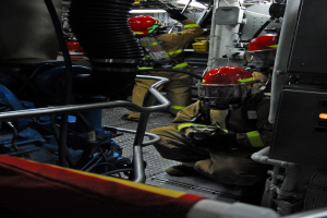 Feuerwehrleute in Helmen und Schutzausrüstung arbeiten an einem Feuerwehrauto mit Rohren und einer Leuchte im Hintergrund.