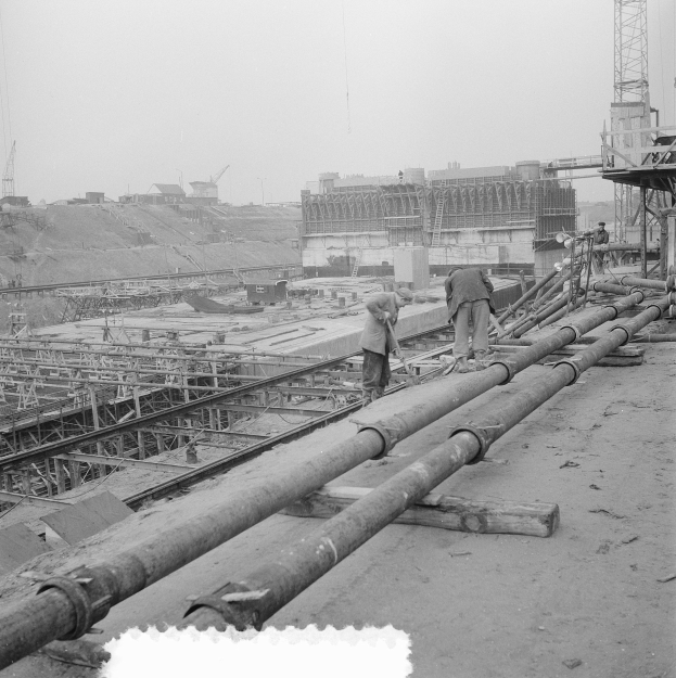 Schwarzes und weißes Foto von Männern auf einer Baustelle mit Rohren im Vordergrund und Gebäuden, Kränen und Himmel im Hintergrund.