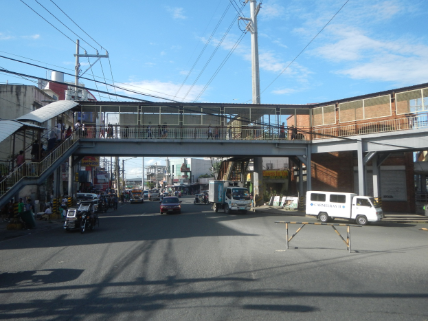 Stadtstraße mit fahrenden Fahrzeugen, eine Fußgängerbrücke mit Menschen, Strommasten mit Drähten, Gebäude und ein bewölkter Himmel.