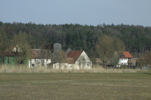 Ein altes Farmhaus steht in einer grünen Wiese umgeben von Bäumen, mit Häusern und einem klaren blauen Himmel im Hintergrund.