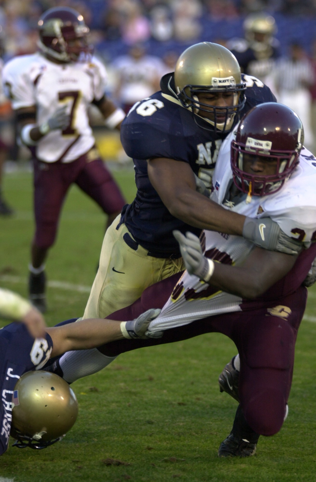 Zwei Fußballspieler in Helmen und Uniformen kämpfen auf dem Feld gegeneinander, während Teamkollegen und Zuschauer im Hintergrund beobachten.