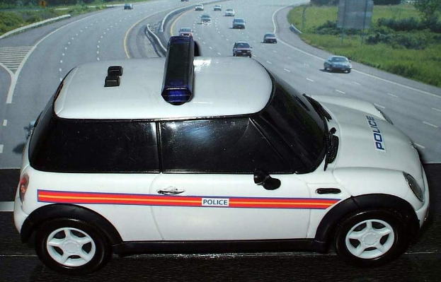 Weißes Polizeiauto mit "Polizei"-Aufschrift auf einer Straße vor einer Stadt, umgeben von fahrendem Verkehr und grasigen Bereichen mit Bäumen auf beiden Seiten.