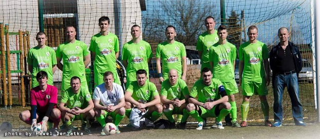 Gruppe von Männern in grünen T-Shirts, die auf einem Fußballfeld stehen und knien, mit einem Ball und einem Torpfosten umgeben von Bäumen, Gebäuden und Fahrzeugen.