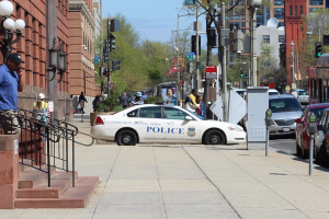 Eine belebte Stadtstraße mit einem parkenden Polizeiwagen, fahrende Fahrzeuge einschließlich eines Busses und eines Autos, Fußgänger auf dem Gehweg, Gebäude mit Fenstern und Geländern, Laternenpfähle, Bäume und einen klaren blauen Himmel im Hintergrund.