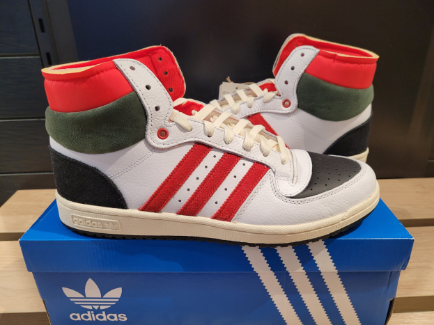 Weiße, rote, grüne und schwarze Adidas Originals Top Ten Hi-Schuhe auf einer blauen und braunen Kartonbox, die auf einem cremefarbenen Tisch mit einer schwarzen Wand im Hintergrund steht.