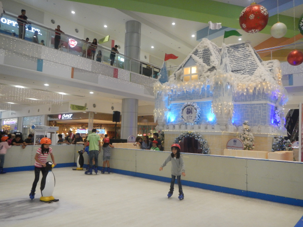 Menschen auf einem Eislaufplatz in einer Mall umgeben von festlichen Dekorationen, mit anderen auf Geländern und Säulen.