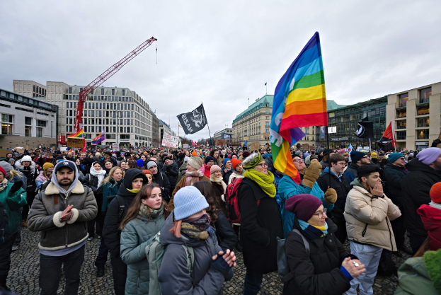 Eine große Gruppe von Menschen steht vor einem Gebäude, hält Fahnen und Transparente mit der Aufschrift "Lgbtq+ rights march in Berlin", einige tragen Mützen und Taschen, mit Gebäuden, einem Kran und Wolken im Hintergrund.