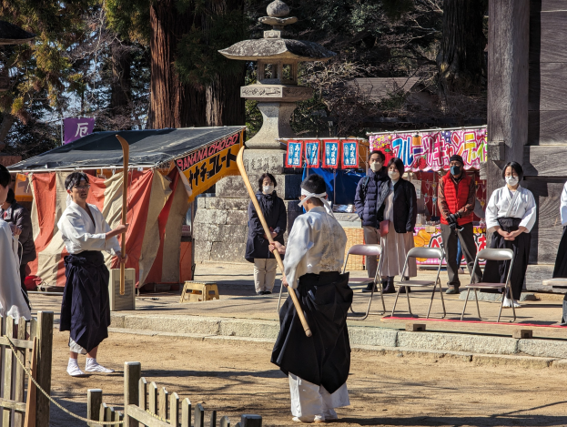 Eine Gruppe von Menschen in traditioneller Kleidung versammelt sich im Freien in Kyoto, einige tragen Masken und halten Holzstöcke, während Stühle, Banner und ein Zelt gegen einen klaren blauen Himmel zu sehen sind.