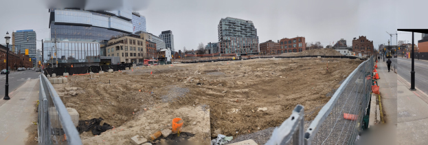 Baustelle mit einem großen Haufen Erde, umgeben von Gebäuden, Straßeninfrastruktur, Fahrzeugen, Fußgängern, Bäumen und einem klaren blauen Himmel.