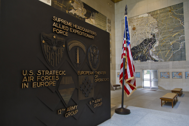 Der Eingang zum U.S. Strategic Air Forces Headquarters in Europa, mit einer amerikanischen Flagge, Bänken, einer Texttafel, Fotorahmen und einer beleuchteten Decke mit einer Tür im Hintergrund.