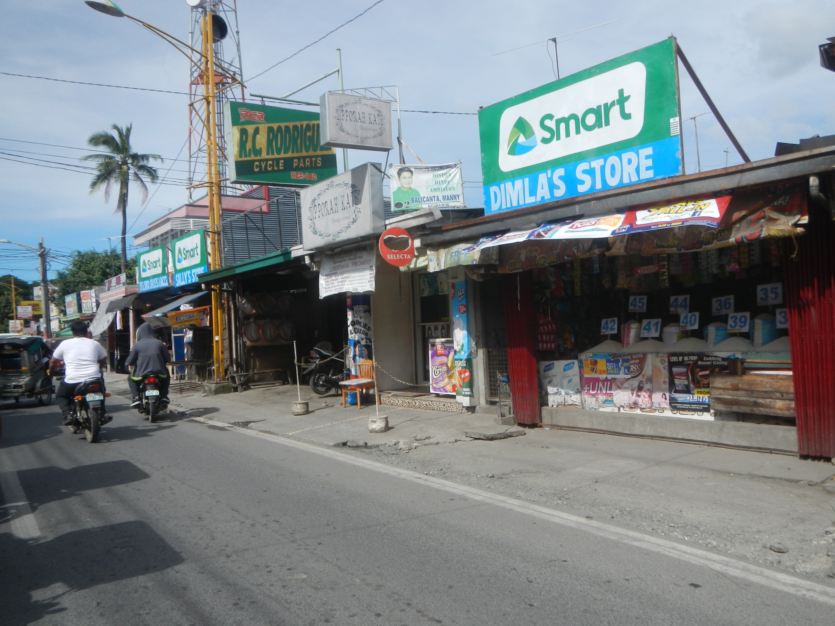 Eine belebte Straße mit Motorrädern, Autos, Gebäuden, Schildern, Strommasten, Kabeln, Straßenlaternen, Bäumen und einem bewölkten Himmel.