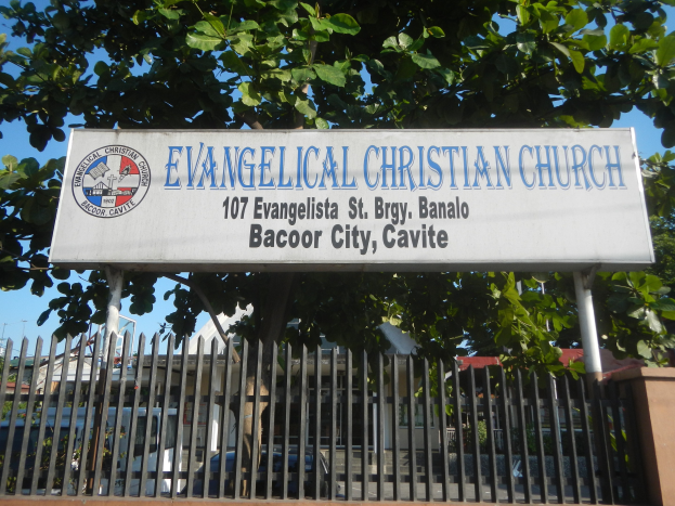 Eine evangelische Kirche in der Stadt Bacoor, Cavite, mit einem Schild, einem Metallzaun, einer Wand, Bäumen, Fahrzeugen, Gebäuden, Polen und einem bewölkten Himmel.