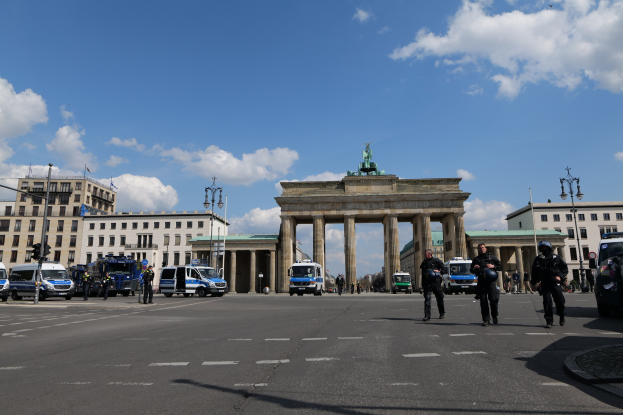 Polizisten vor dem Brandenburger Tor in Berlin, Deutschland, mit Säulen und Statue des Tores, umgeben von Gebäuden, Fahrzeugen, Laternen und Verkehrszeichen unter einem bewölkten Himmel.