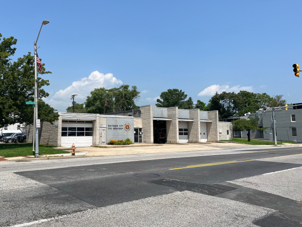 Eine Feuerwache an einer Straßenecke mit umliegenden Gebäuden, Fahrzeugen, Verkehrszeichen, einer Hydranten, Grünfläche und einem bewölkten Himmel.