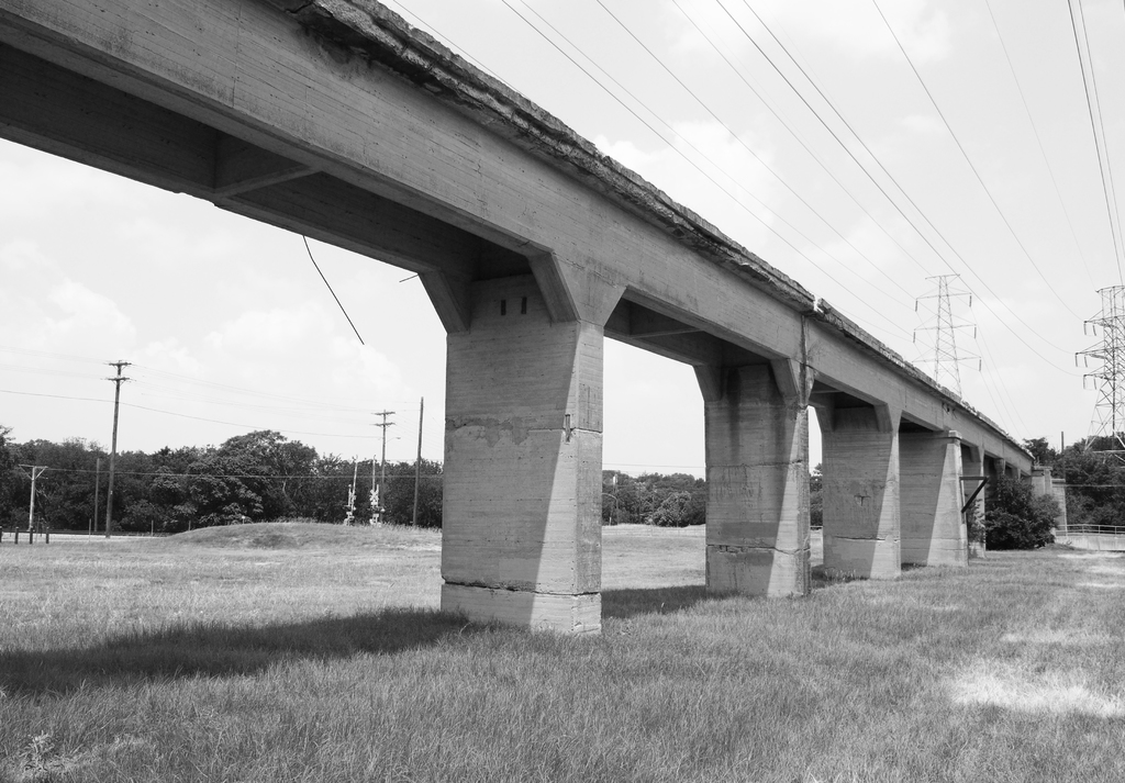 Eine Betonbrücke auf einer grünen Wiese mit einem großen Strommast und Kabeln darüber, umgeben von Bäumen und zusätzlichen Strommasten im Hintergrund.
