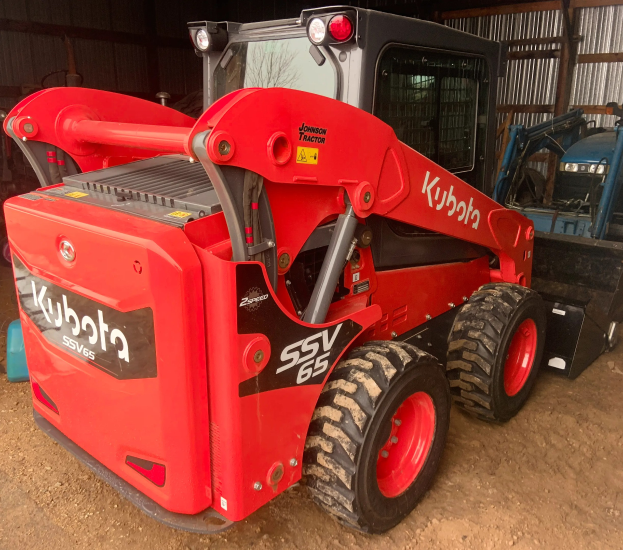 Roter Kubota SSV65 Radlader vor einer Scheune geparkt mit anderen Fahrzeugen im Hintergrund.