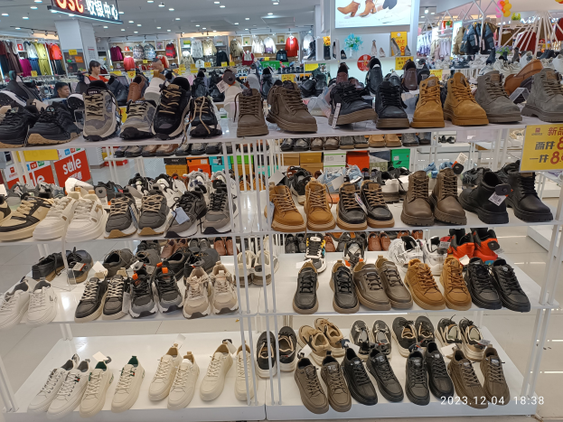 Ein Schuhgeschäfts-Innenraum mit verschiedenen Schuharten auf Gestellen, Preisetiketten sichtbar, Menschen im Hintergrund, Kleiderständer, Deckenlampen, ein Bildschirm und ein Wasserzeichen, das einen Standort in China angibt.