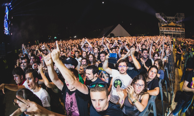 Menschenmenge auf einem Musikfestival mit erhobenen Händen, sitzend und stehend, einem Metallzaun auf der rechten Seite und Gebäuden mit Lichtern im Hintergrund unter einem dunklen Himmel.