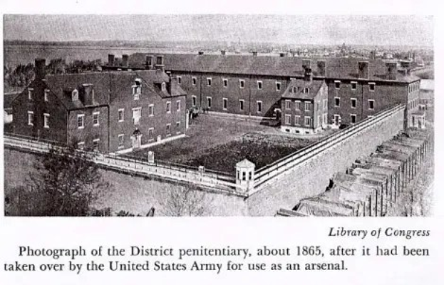 Schwarzes und weißes Foto des Bezirksgefängnisses aus dem Jahr 1865, das als U.S.-Armee-Lager genutzt wurde, zeigt Gebäude, Fenster, Bäume, eine Wand und Text am unteren Rand.
