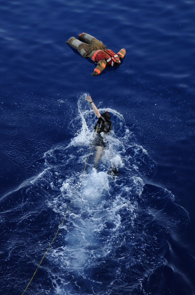 Ein Retter in einem Rettungswesten, der an einem Seil über ruhigem, klarem Wasser unter strahlendem Sonnenschein hängt.