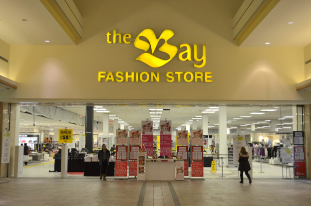 Außenansicht eines Bay Fashion Stores in einem Einkaufszentrum mit Menschen, die gehen, Texttafeln an den Wänden, Säulen, Deckenlampen und Hintergrundauslagen.
